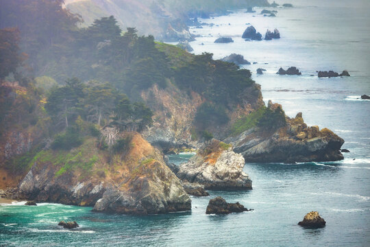 Misty Big Sur coastline along Highway 1 in California. Rugged cliffs covered with green cypress trees rise above turquoise waters with rocky formations extending into the Pacific Ocean hazy horizon - Powered by Adobe