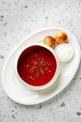 Tomato soup with herbs served in a white bowl on a plate, accompanied by glazed bread rolls and sour cream. Minimalist food presentation on a speckled countertop.