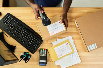 Black man scanning shipping label on cardboard box at desk with computer keyboard, barcode scanner, payment terminal and envelopes, processing packages for delivery or logistics service