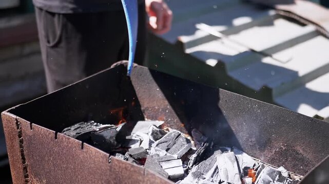 Fanning the embers in a metal brazier using a blue shovel, this shot captures the essence of outdoor cooking preparation. The orange glow of the coals and the gentle smoke evoke the warmth of a summer