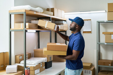 Young adult Black man organizing cardboard boxes on metal shelves in warehouse storage room, standing and reaching for package while wearing blue cap and polo shirt, surrounded by parcels