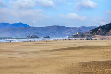 Wide sandy expanse of Ocean Beach in San Francisco with people walking and relaxing along the shoreline. Waves roll in from the Pacific Ocean with hills and the historic Cliff House visible in the