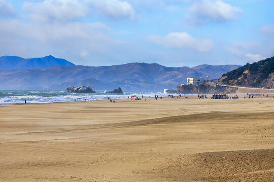 Wide sandy expanse of Ocean Beach in San Francisco with people walking and relaxing along the shoreline. Waves roll in from the Pacific Ocean with hills and the historic Cliff House visible in the
