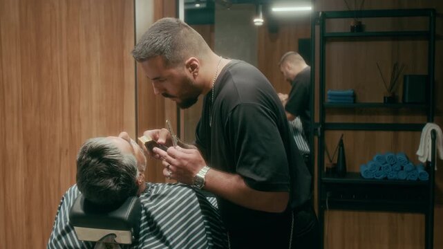 Medium shot of professional young Caucasian barber in black tshirt bending over mature male patron in reclining chair, trimming moustache and beard using electric shaver