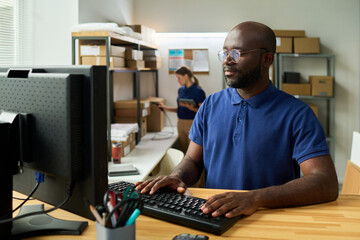 Black man working at desktop computer in warehouse office, typing on keyboard and focusing on screen while young Caucasian woman organizing packages on shelves in background