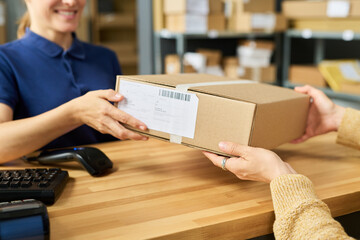 Obraz na płótnie Canvas Caucasian woman handing cardboard package to another woman across wooden counter in shipping or delivery service office, both women interacting with parcel during transaction Obraz na płótnie Canvas Caucasian woman handing cardboard package to another woman across wooden counter in shipping or delivery service office, both women interacting with parcel during transaction