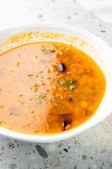 Homemade vegetable soup in white bowl on gray countertop. Rich orange broth with carrots, potatoes, beans, herbs. Healthy, rustic, freshly made meal. Top view, natural light