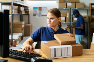 Caucasian young adult woman working at shipping desk holding cardboard boxes and using computer while Black man in background sorting packages in warehouse environment