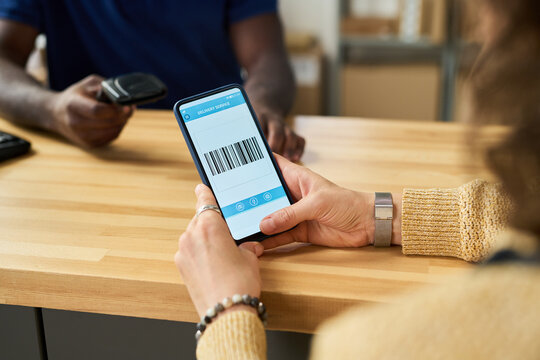 Caucasian young adult woman holding smartphone displaying barcode, while Black man scanning screen with handheld barcode reader across wooden counter in modern retail or shipping setting