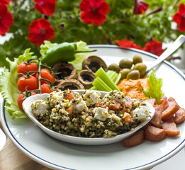 Bulgur Wheat Onion Salad with Feta Cheese, Pepper Dip, Sausages, Tomatoes, Mushrooms, Green Olives, Celery Sticks on White Plate, Garden Petunia Background
