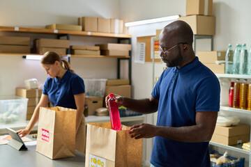 Black middle aged man packing food delivery bag with bottled drink while Caucasian young adult...