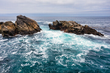 Rocky shoreline along Highway 1 in Big Sur, California, USA. Rugged cliffs meet turquoise Pacific waves, with coastal mountains rising in the background, showcasing iconic scenery of the Pacific Coast