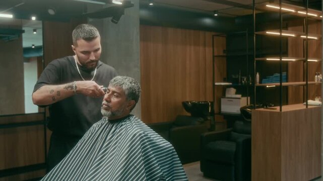 Medium full shot of young Caucasian barber in black clothes trimming hair on temples using clipper for mature Indian client, while working on stylish crew cut at barbershop