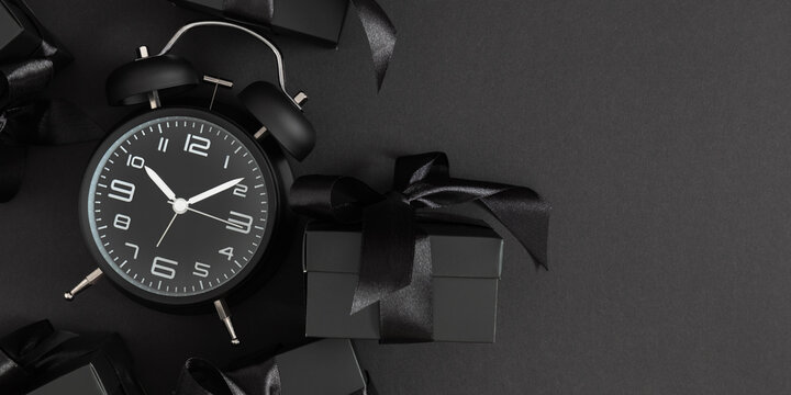 Festive black background dedicated to Black Friday. Featuring a black alarm clock and gift boxes on a black surface.