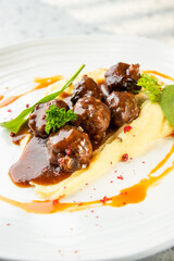 Meatballs in brown sauce served over mashed potatoes, garnished with spinach and parsley, plated on white dish with decorative drizzle. Gourmet presentation on light background.
