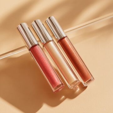 Tre tubi di lip gloss nude su bacchetta di vetro, sfondo beige caldo e ombre soft. Composizione diagonale, look pulito per cataloghi, campagne social e shop online.