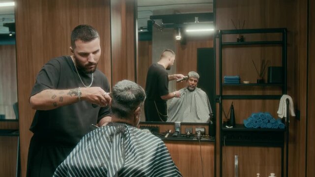Medium shot of young Caucasian male hairdresser in black tshirt, with tattoos working on buzz or crew cut with trimmer for middle-aged Asian man, sitting in stripy cape in chair in front of mirror
