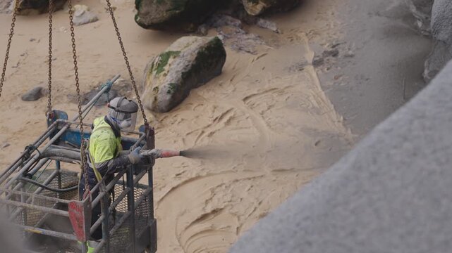 Construction worker applies concrete using shotcrete technique to stabilize coastal cliff for erosion protection and shoreline reinforcement. Safety cage and protective gear ensure secure operation