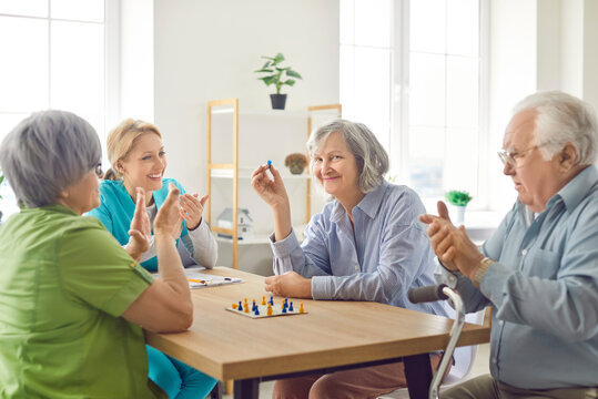 Happy caregiver engaged in entertainment with senior patients, sitting at table and playing board games together at a nursing home. People companionship and leisure time activities in elderly care.