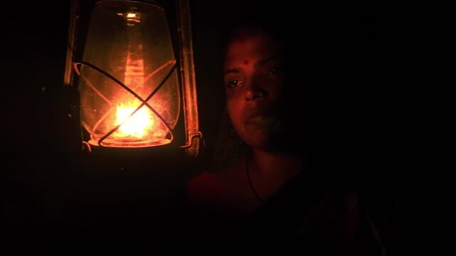 A calm, thoughtful Indian woman holds a glowing kerosene lantern in the warm darkness, her pensive expression evoking mystery, hope, and quiet strength as the soft light illuminates the night.