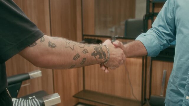 Medium shot of hands of happy unrecognizable customer in blue casual shirt thanking tattooed barber for haircut and good service with handshake after appointment at hair salon