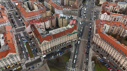 Obraz premium Top-down view of converging boulevards with a tram corridor, angled corner block and red-tiled facades; zebra crossings, parking rows and queuing traffic animate a dense winter streetscape under overc