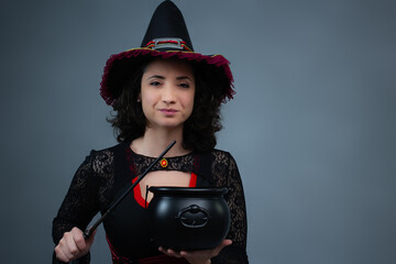 Happy mid-adult witch holding a cauldron and magic wand, looking at camera. Ideal for Halloween ads, trick or treat, magical spells, and fairy tale villain. Horizontal studio portrait with copy space.