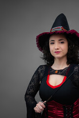 Charming mid-adult witch holding a magic wand, looking at camera. Ideal for Halloween ads, trick or treat, magical spells, and fairy tale villain. Vertical studio portrait with copy space.