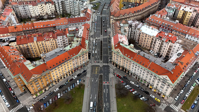 Top-down aerial of a symmetrical crossroads: red-tiled historic blocks form a sharp V, boulevards with tram lines, crosswalks and traffic, parked cars along facades; winter light and overcast mood. - Powered by Adobe
