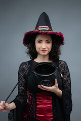 Charming mid-adult witch pointing a magic wand at empty cauldron, looking at camera. Perfect for Halloween, witches brew, and fairy tale villain. Studio portrait with copy space on gray background.