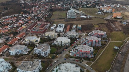 Aerial view of a suburban neighborhood with low-rise apartment blocks, colorful facades, curved streets and parked cars, bordered by open fields and a highway; winter light and bare trees.