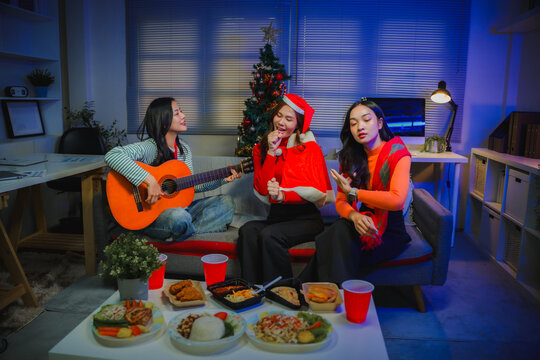 Three asian women friends celebrating Christmas holiday, singing, playing guitar, and enjoying food at home party