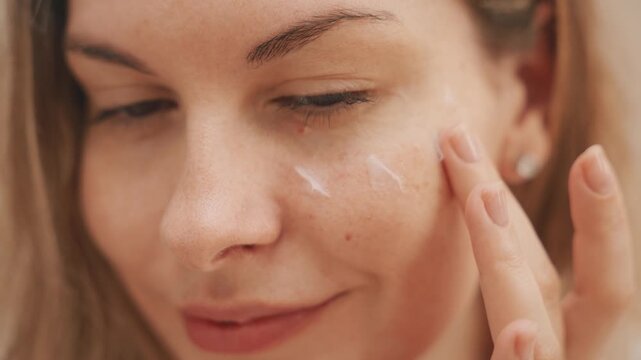 Young adult happy woman applying cream under eyes on face healthy skin care moisturizing nutrition white gel. girl looking in mirror reflection uses product natural cosmetics anti-aging rejuvenation