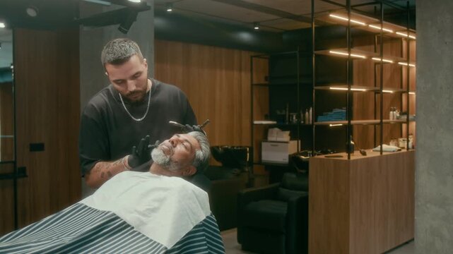 Medium shot of young Caucasian barber in black tshirt and gloves applying lather or foam to Indian male clients face before shaving with straight razor blade during indulgent grooming treatment