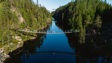 Explore the stunning natural beauty of Julma Olkky Canyon in Hossa National Park Finland. Visitors walk across a suspension bridge, surrounded by lush forests and reflective waters on a sunny day.
