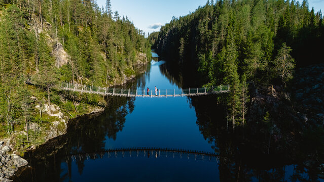 Explore the stunning natural beauty of Julma Olkky Canyon in Hossa National Park Finland. Visitors walk across a suspension bridge, surrounded by lush forests and reflective waters on a sunny day. - Powered by Adobe