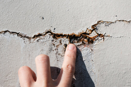 Person inspecting crack in wall with water seeping out. Close up of hand examining  vertical crack in outdoor wall with rust and efflorescence. Repair foundation problems. Selective focus.