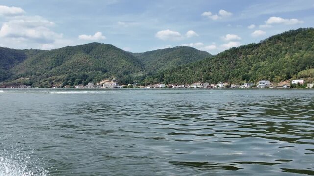 Speedboat navigating the Danube river through the Iron Gates gorge, with forested mountains rising steeply from the water and a village scattered along the bank