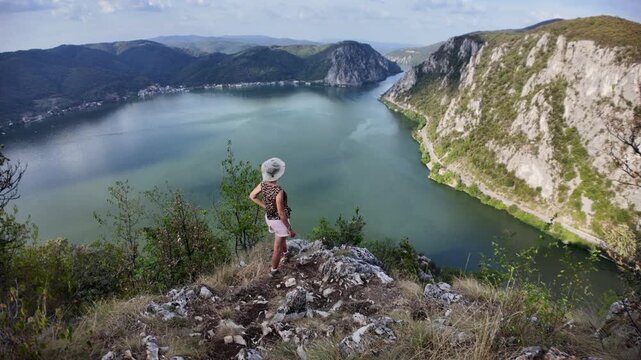 Woman stands on rocky cliff overlooking sweeping Danube gorge, lush green mountains on Serbia-Romania border, enjoying panoramic river landscape and tranquil wilderness vista