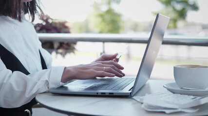 A woman is engaged in remote work using her laptop while seated comfortably at a cozy cafe table, sipping coffee - Powered by Adobe