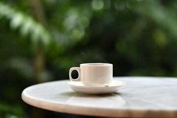 White coffee cup on saucer steaming on round marble table in outdoor garden setting, calm morning atmosphere, shallow depth of field, natural light