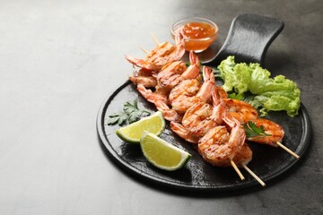 Skewers with tasty grilled shrimps, lime slices, parsley, lettuce and sauce on grey table, space for text. Delicious seafood dish