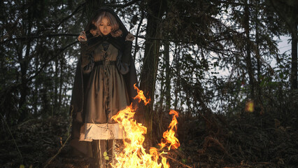 Enchanting hooded woman casts spells by bonfire in mysterious forest, evoking fantasy and magic,...