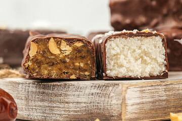 Assorted homemade chocolate-covered protein bars with peanut and coconut filling, cut in half to show texture, arranged on a rustic wooden board with dates and peanuts