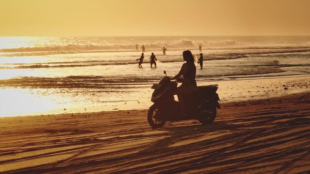 Silhouette of beautiful woman riding scooter along sandy beach, golden warm sunset over ocean waves in Bali, local fishermen fishing in background. Remote wild nature paradise, exotic summer travel - Powered by Adobe