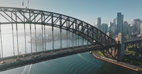 Australia, Sydney: Aerial view of Sydney Harbour Bridge, Sydney Opera House and city skyline in background. Sunny morning light, train and cars crossing the bridge. Modern architecture. Drone