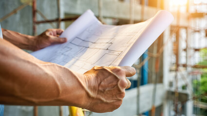 Close-up of a construction worker’s hands holding and examining detailed blueprints on a building site, with scaffolding and unfinished structure visible in the background under warm sunlight