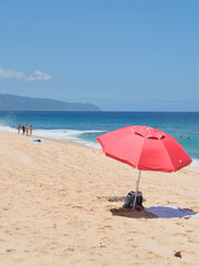Red beach umbrella on a sandy tropical shore with  waves and blue sky. People walk along the coastline in the distance, perfect summer vacation and relaxation scene Hawaii.