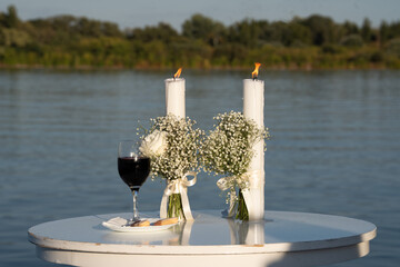 Two religious wedding candles placed on a wooden table beside a glass of red wine on a pier, with a serene lake in the background. Ideal for wedding, romance, or outdoor celebration themes