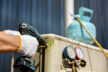 Close-up of an air conditioner technician repairing an outdoor AC compressor with a refrigerant tank and pressure gauge using an electric drill.